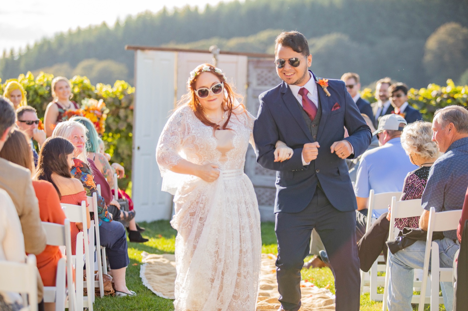 Bride and groom in sunglasses dance down the aisle past seated guests at an outdoor wedding ceremony