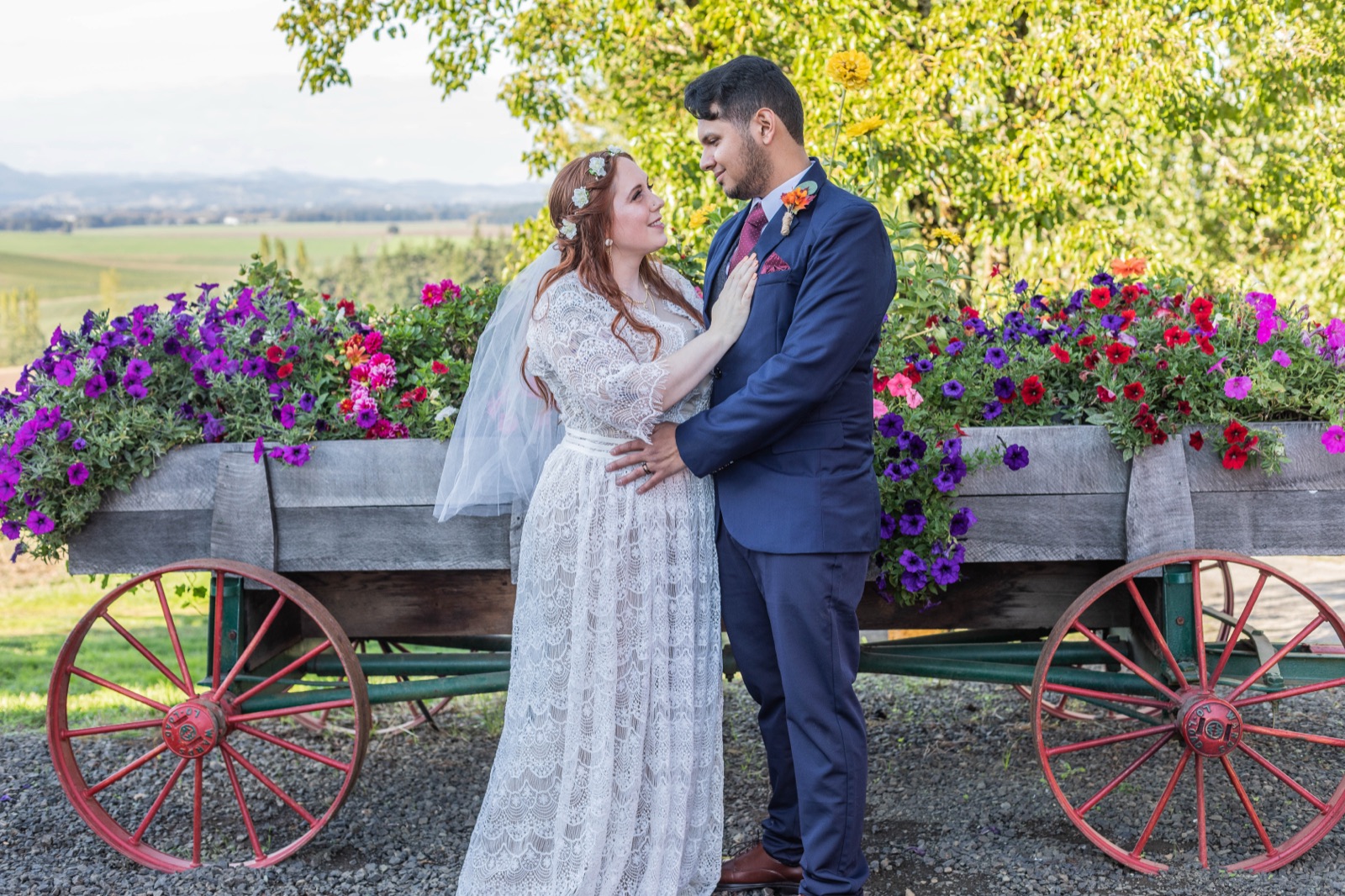 Bride and groom face each other in front of an antique wagon overflowing with purple and red petunias
