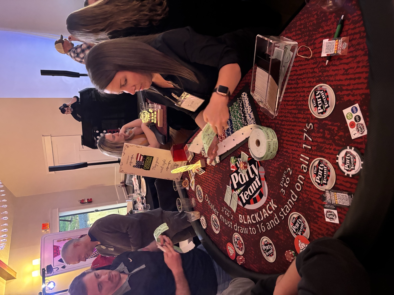 Dealer at a blackjack table hands out raffle tickets while guests play at a casino party, with the Apogee DJ booth in the background