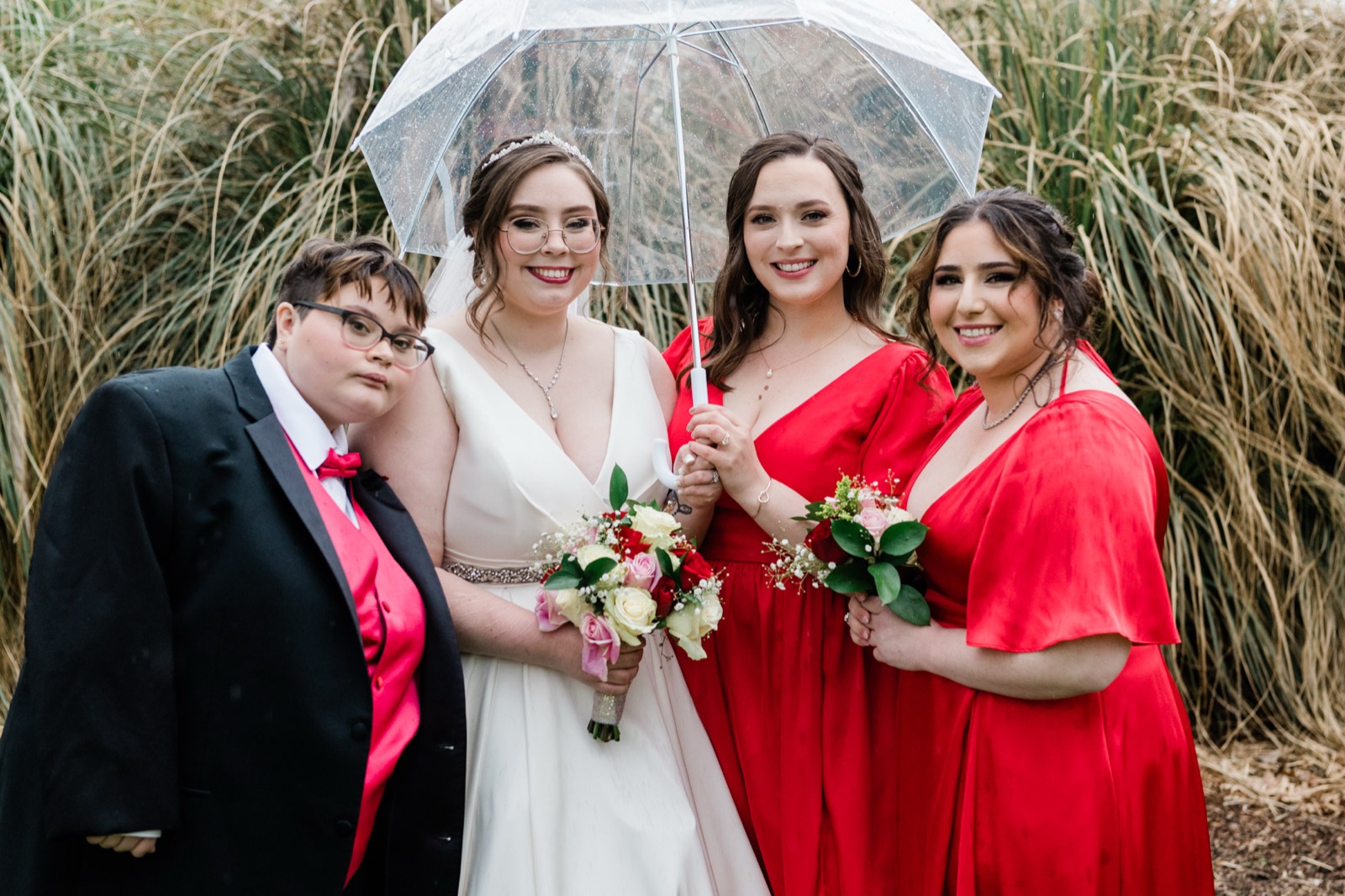 Bride with bridesmaids in red under a clear umbrella in front of tall grass at Green Villa Barn in Independence, Oregon
