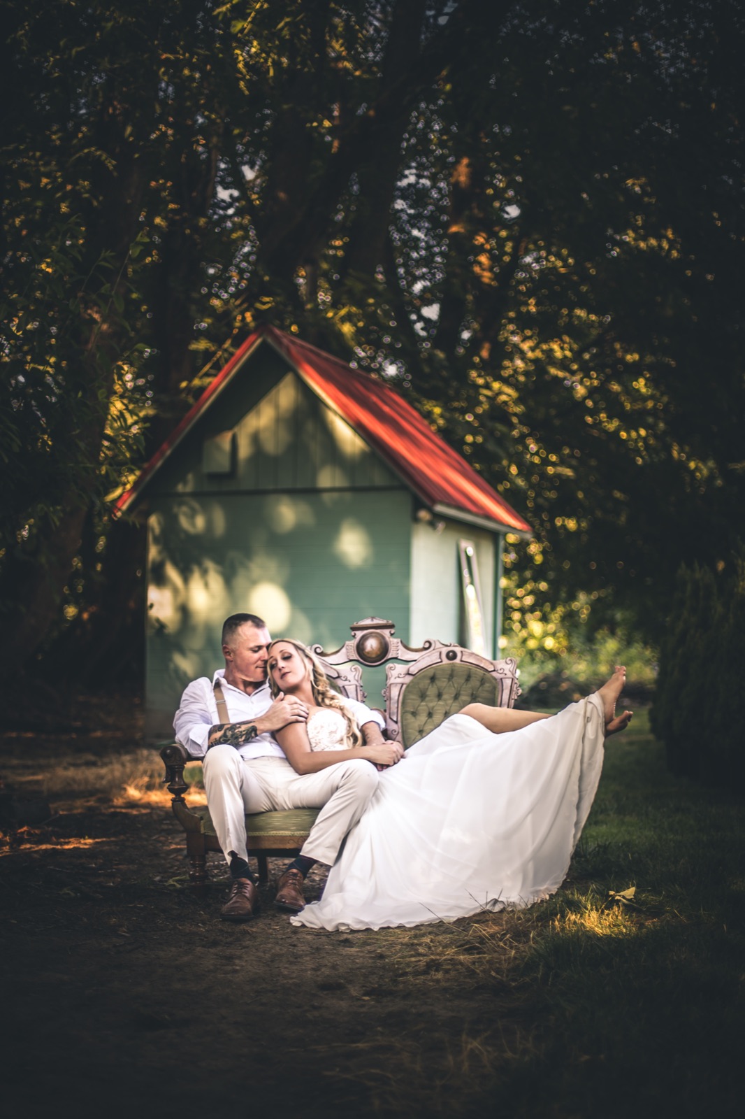 Bride and groom relax on a vintage green settee in front of a small red-roofed shed under trees at golden hour