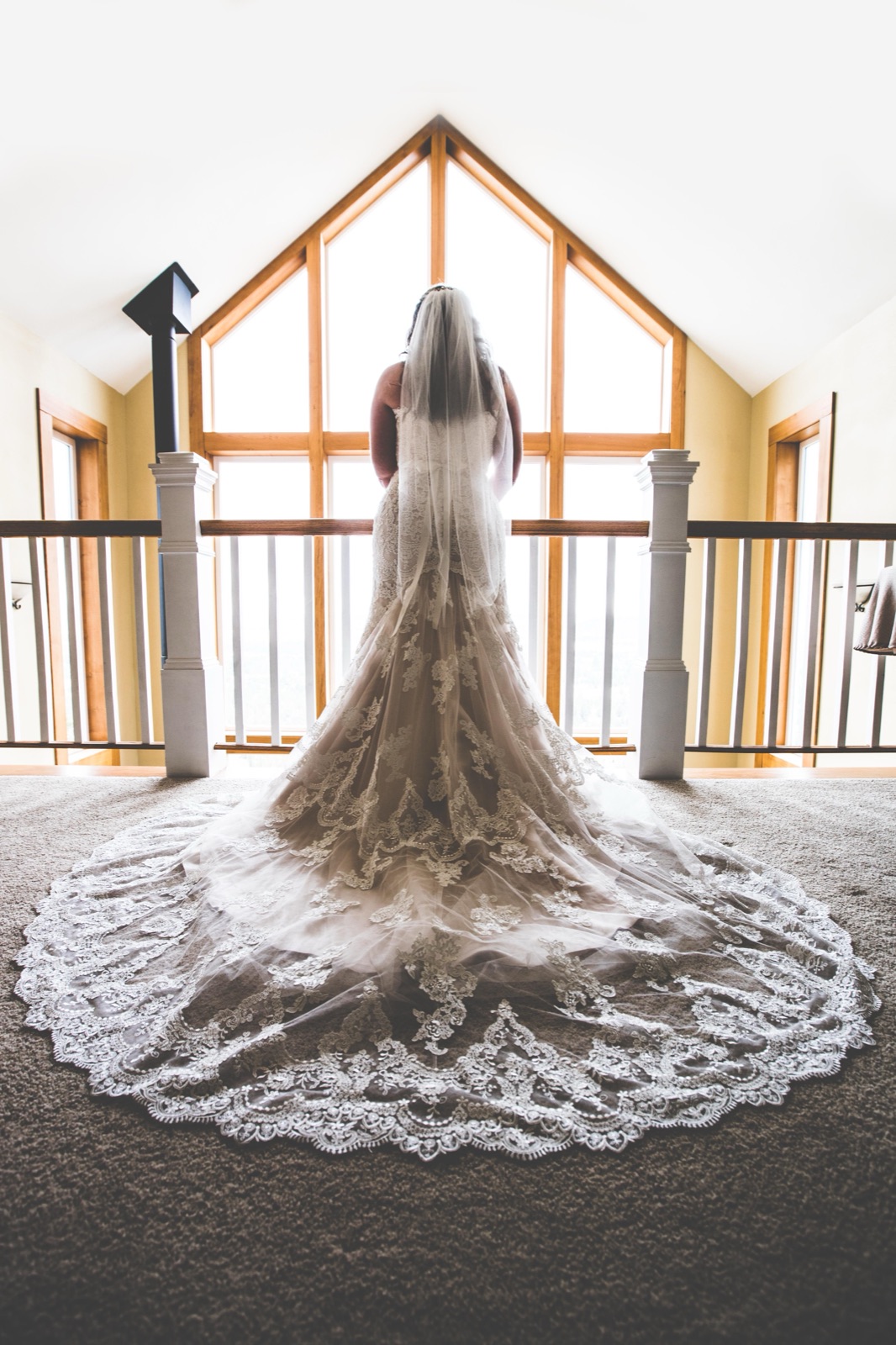 Bride seen from behind at a bright peaked window, her lace dress train and veil spread across the carpet
