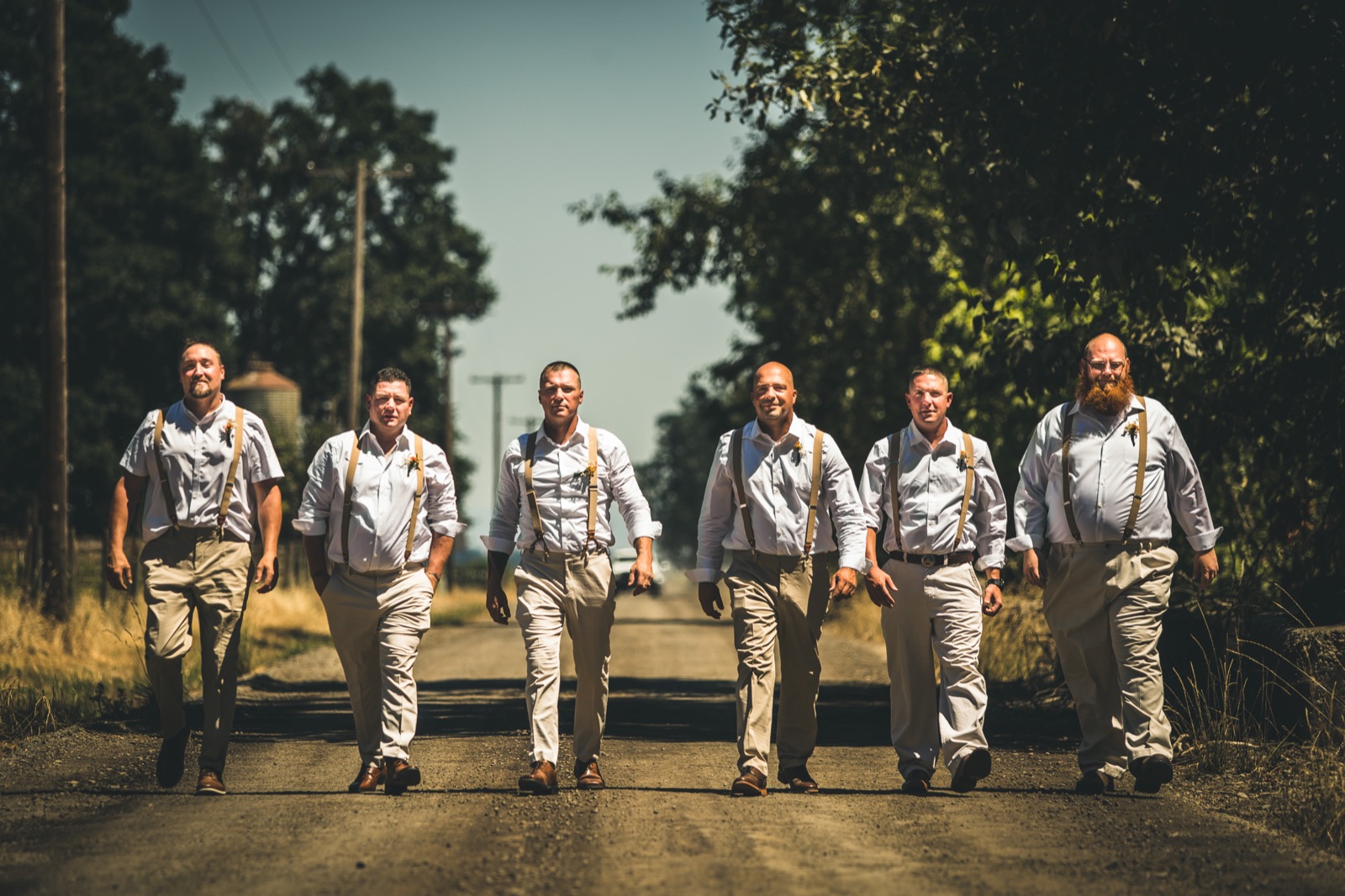 Six groomsmen in white shirts, tan suspenders, and khakis walk down a gravel country road