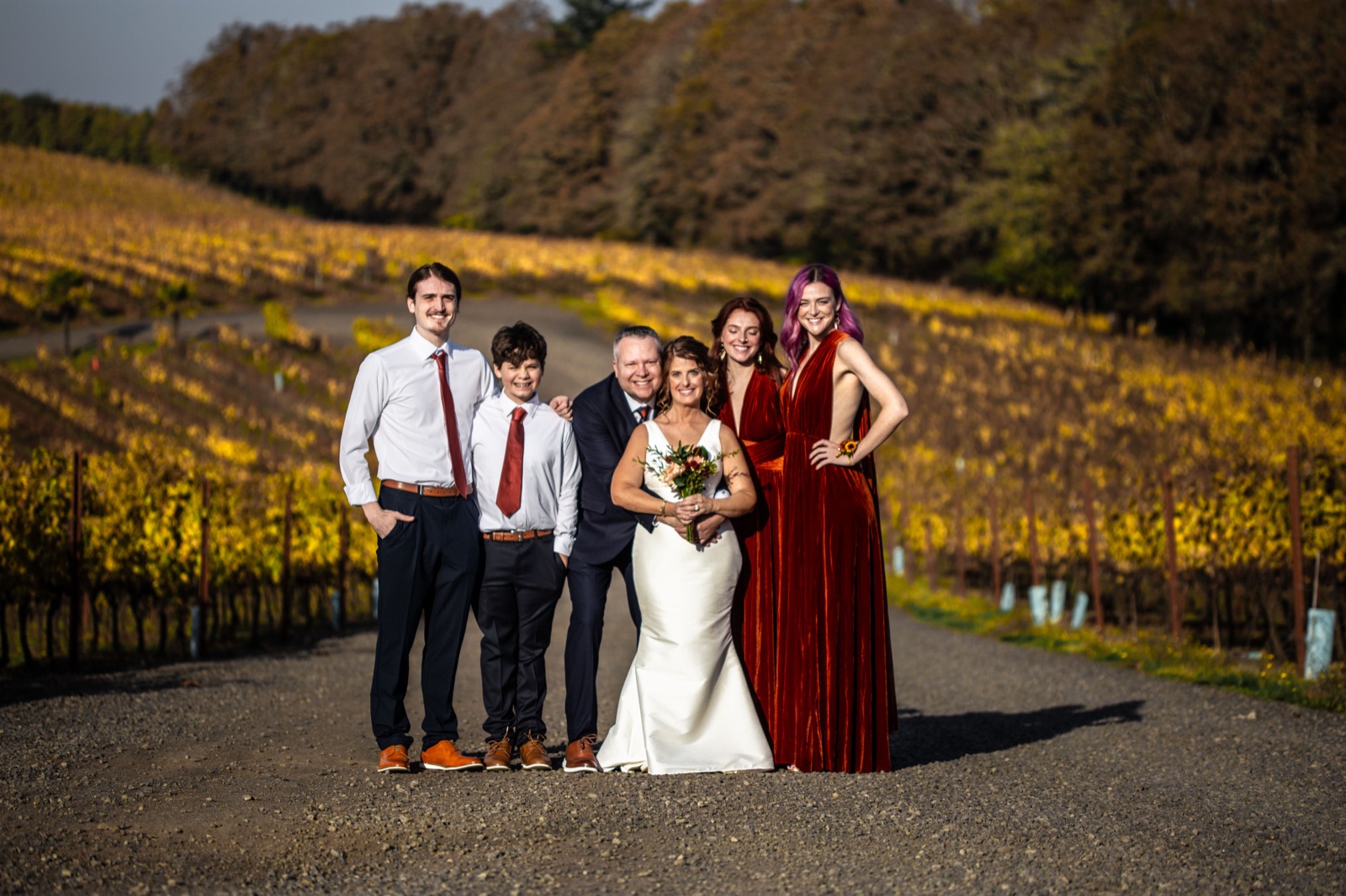 Bride and family in navy suits and rust velvet dresses posing on a gravel road between autumn vineyard rows