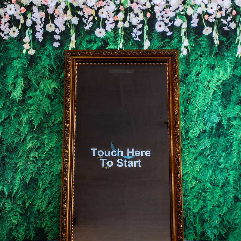 Gold-framed Magic Mirror photo booth in front of a greenery and flower backdrop