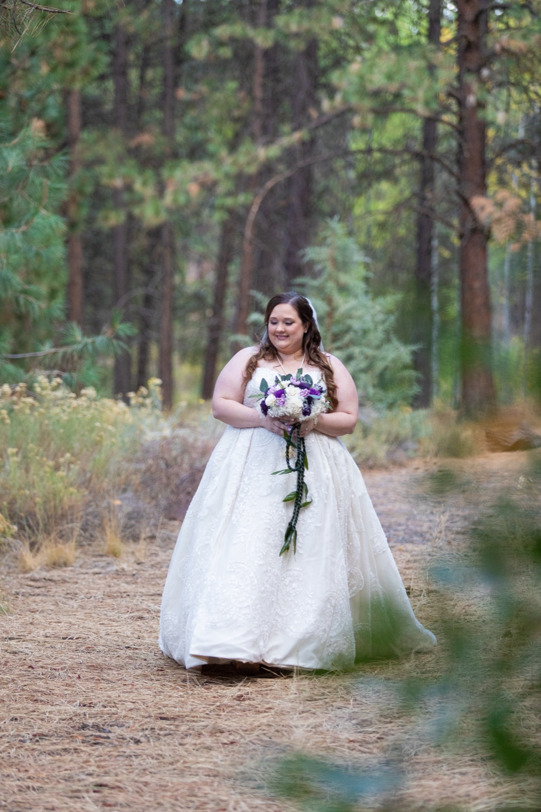 Bride in a lace ball gown holding a purple and white bouquet, walking on a pine-forest path