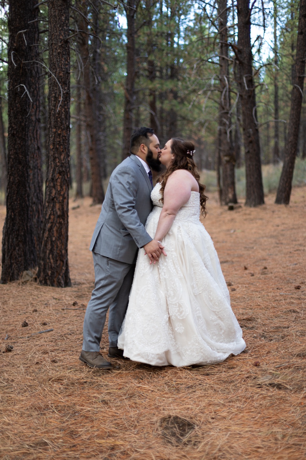 Bride and groom kiss holding hands among tall ponderosa pines, her lace gown against a pine-needle floor