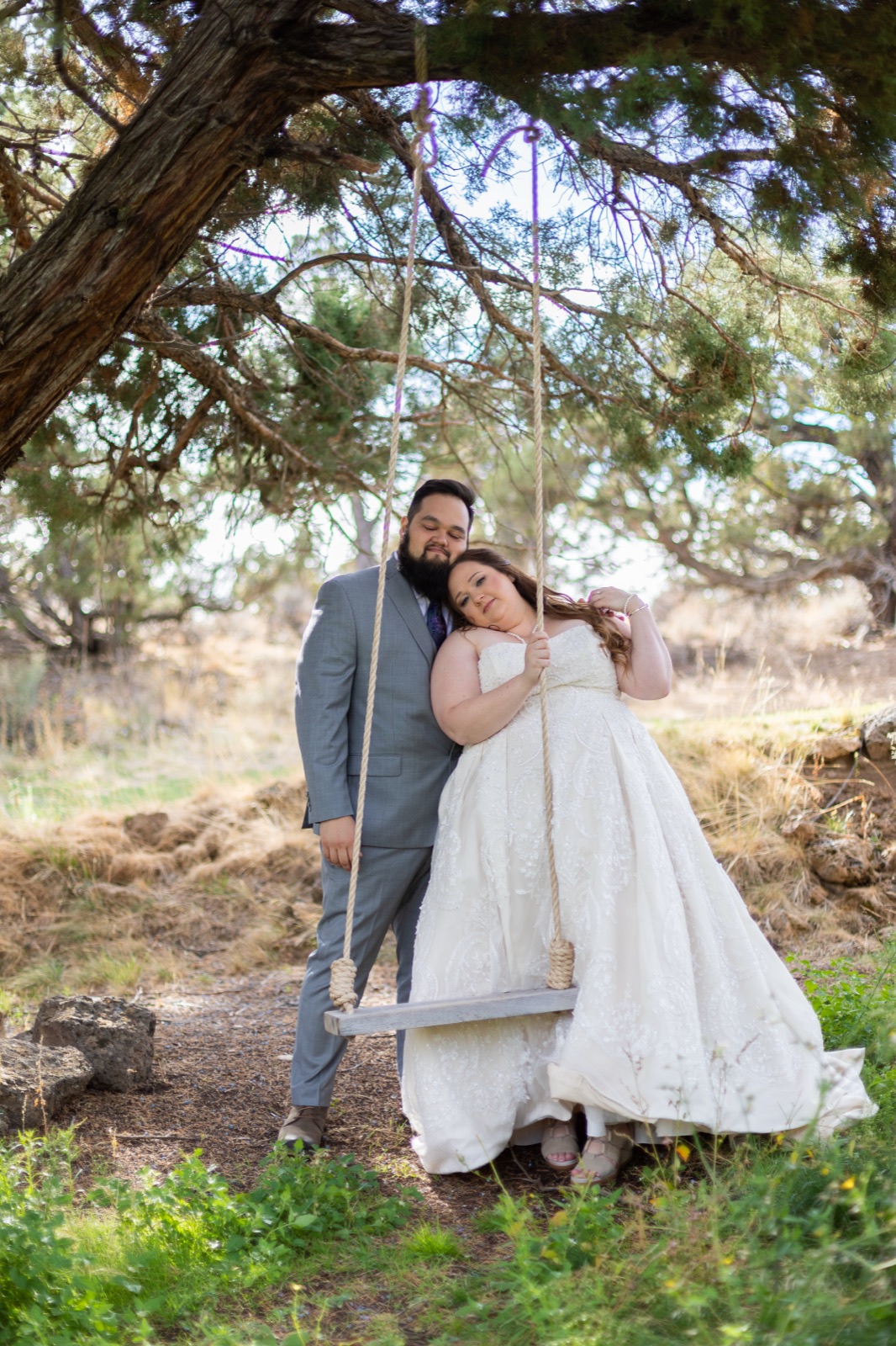 Bride sits on a rope tree swing leaning her head against the groom standing beside her