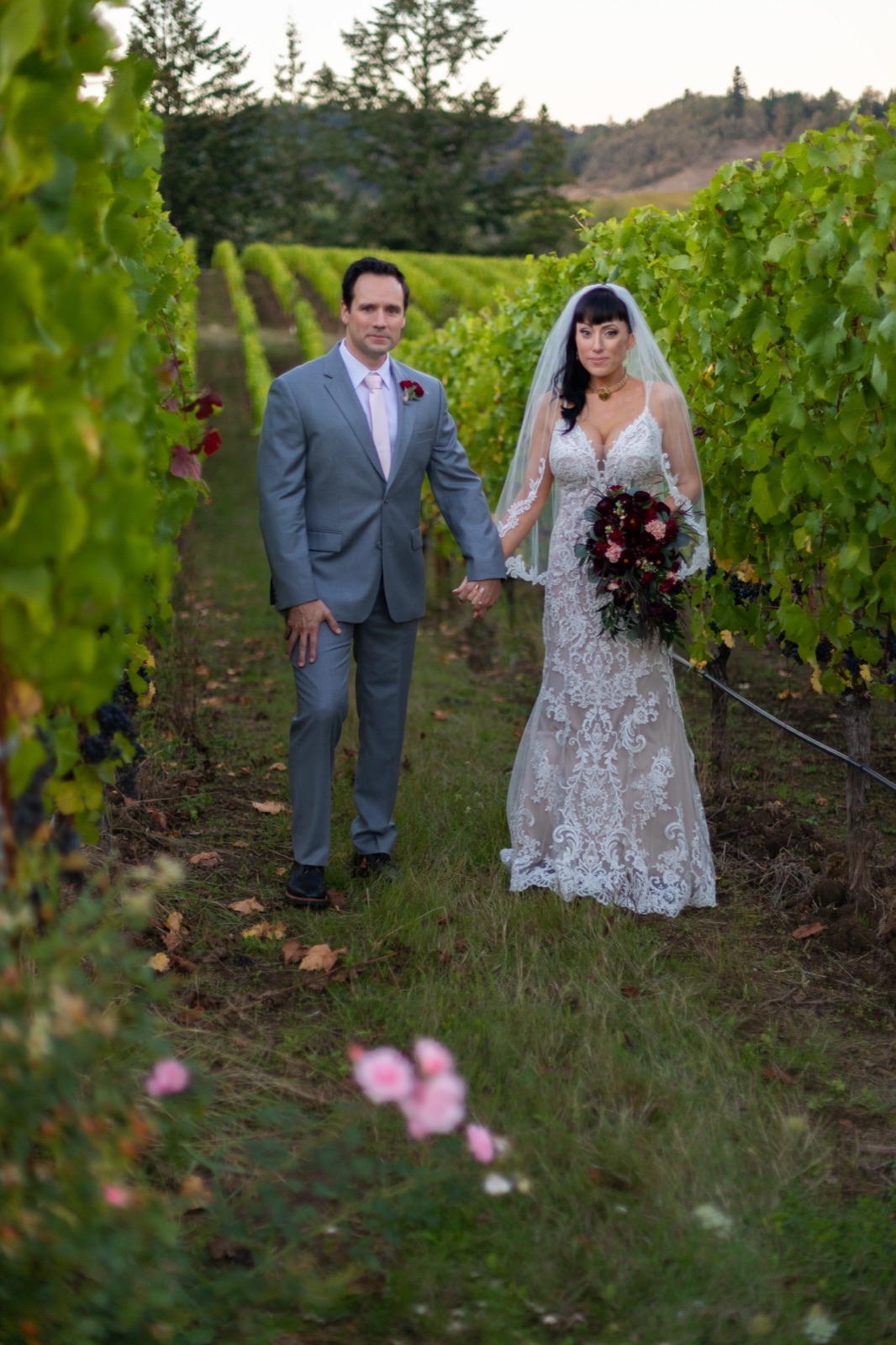 Bride and groom hold hands between rows of grapevines, bride carrying a dark red bouquet