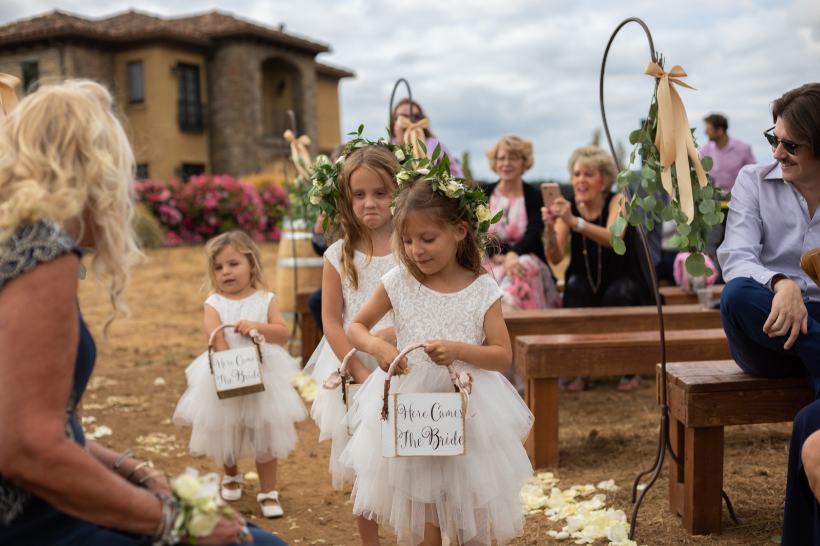 Three flower girls in white tulle dresses and flower crowns carry 'Here Comes The Bride' baskets down an outdoor aisle