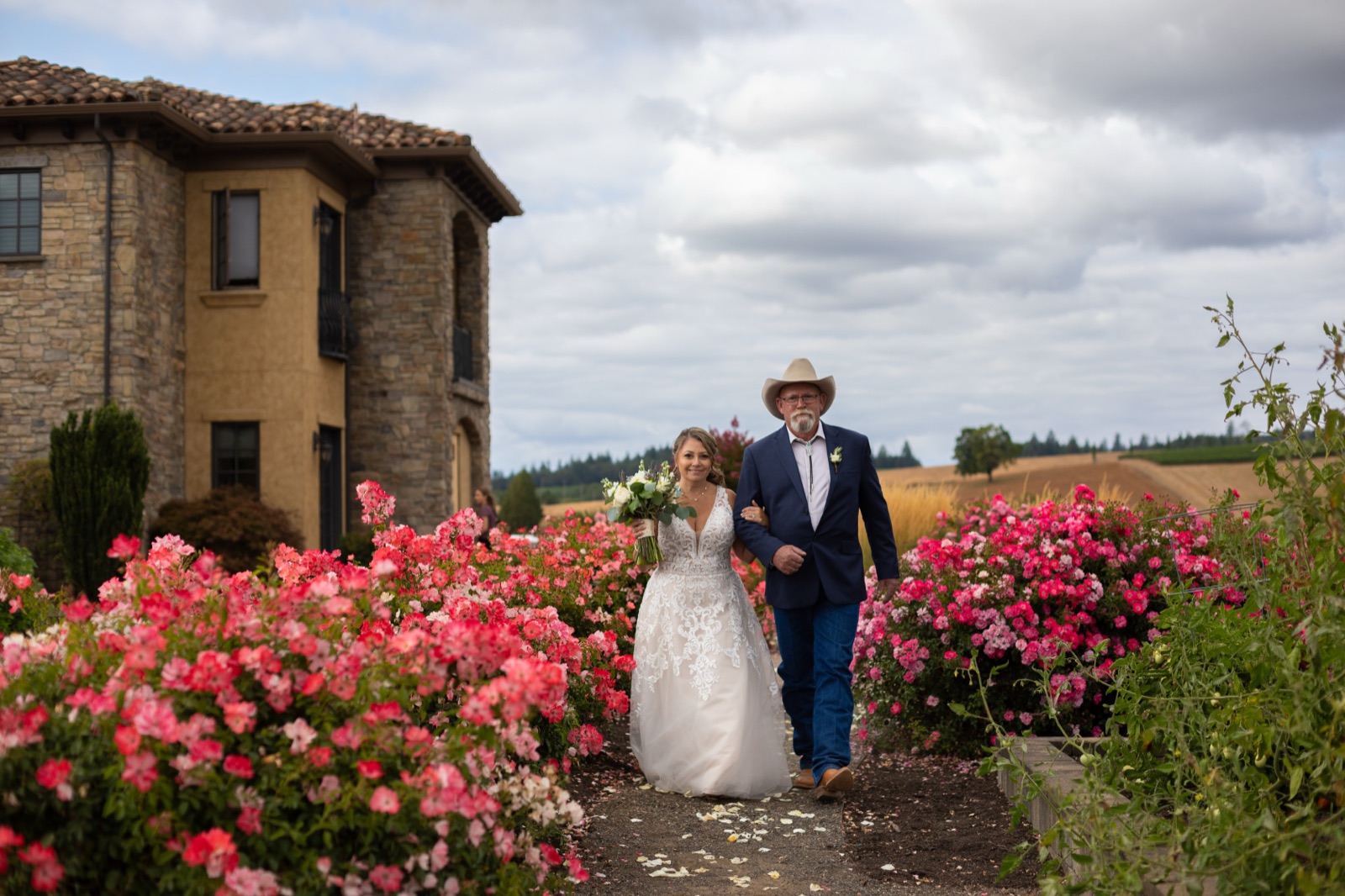Bride with bouquet walks arm in arm with a man in a cowboy hat and navy suit down a path lined with pink roses