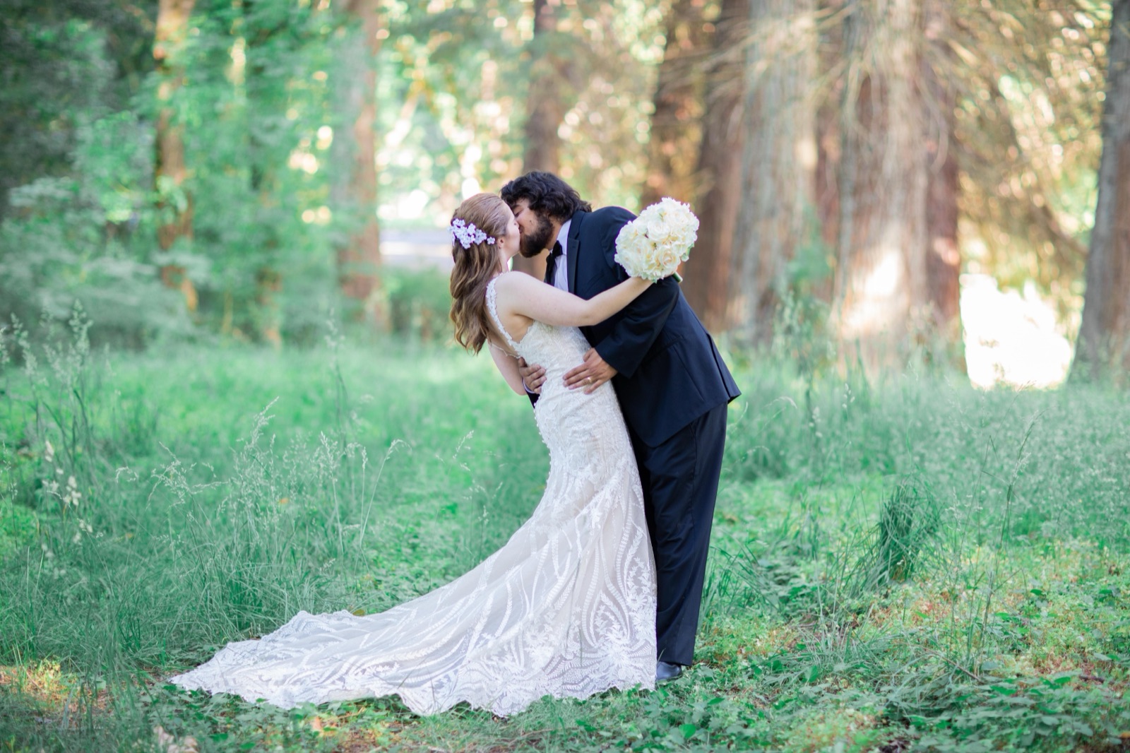 Groom kisses the bride while holding her bouquet aloft in a sunlit meadow surrounded by tall trees
