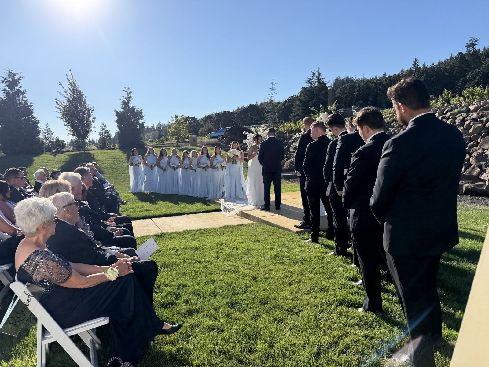 Hillside vineyard wedding ceremony at West Hills Vineyards near Salem, Oregon, full wedding party in white and black under a clear sky