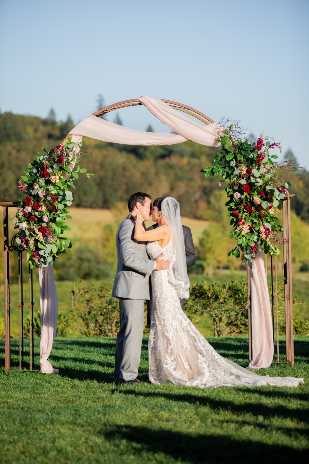 Bride and groom kiss under a round metal arch with greenery, red flowers, and blush drapery on a lawn at Zenith Vineyard