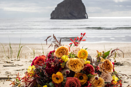 A vibrant floral bouquet on a sandy beach. in the background is photographed the ocean and Oregon's Iconic Haystack Rock at Pacific City, Oregon