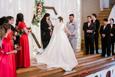 A couple getting married at Green Villa Barn. The groomsmen on the right are wearing black. The bridesmaids are wearing a vibrant red.