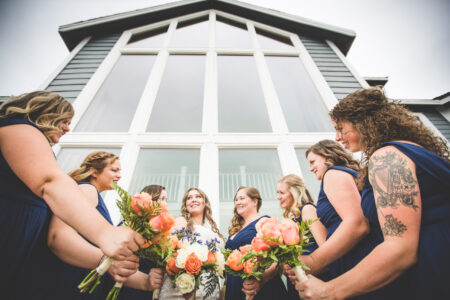 A bride and her bridesmaids show off their bouquets in front of a building with very tall windows.