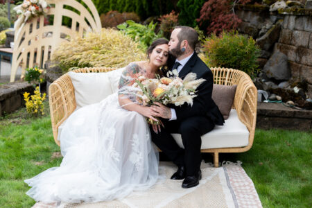 Bride and groom sitting on rattan loveseat with boho floral styling Oregon outdoor wedding portrait